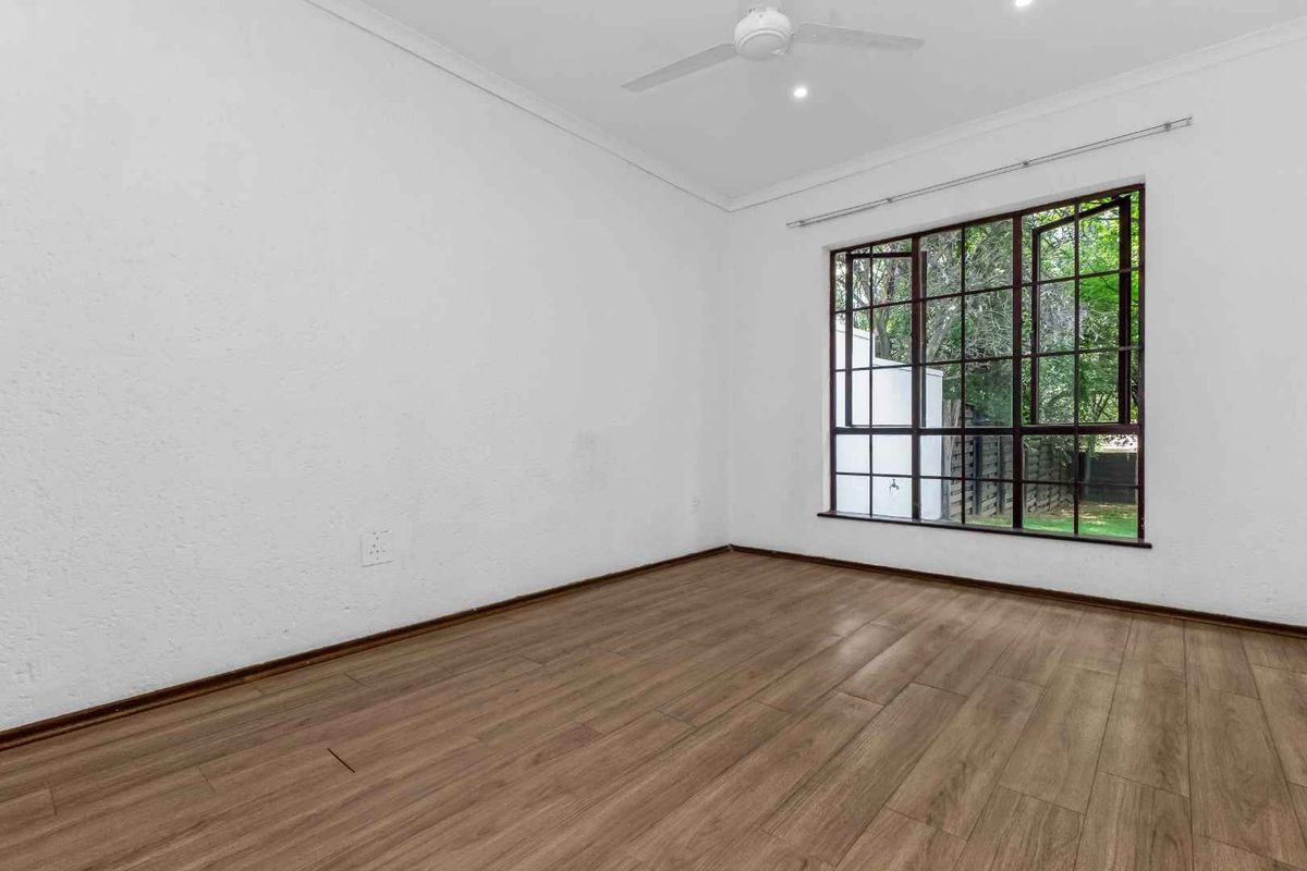 A bright, empty bedroom featuring modern laminate flooring, a ceiling fan, and a large window overlooking the garden.