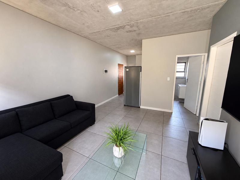 Open-plan living area featuring large format floor tiles and an exposed concrete ceiling with recessed lighting, leading to adjacent rooms.