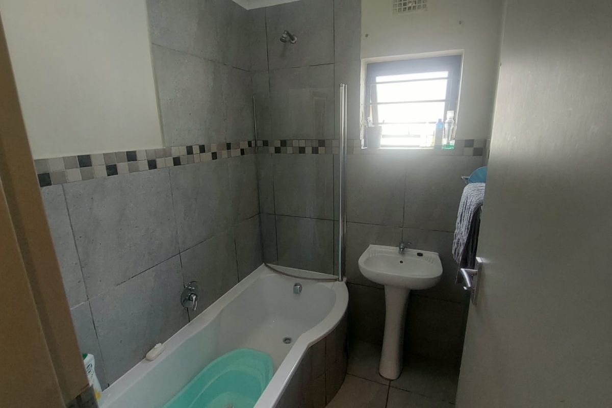 A functional bathroom featuring a bathtub with a glass shower screen, a pedestal basin, and grey wall tiling with a decorative mosaic border.