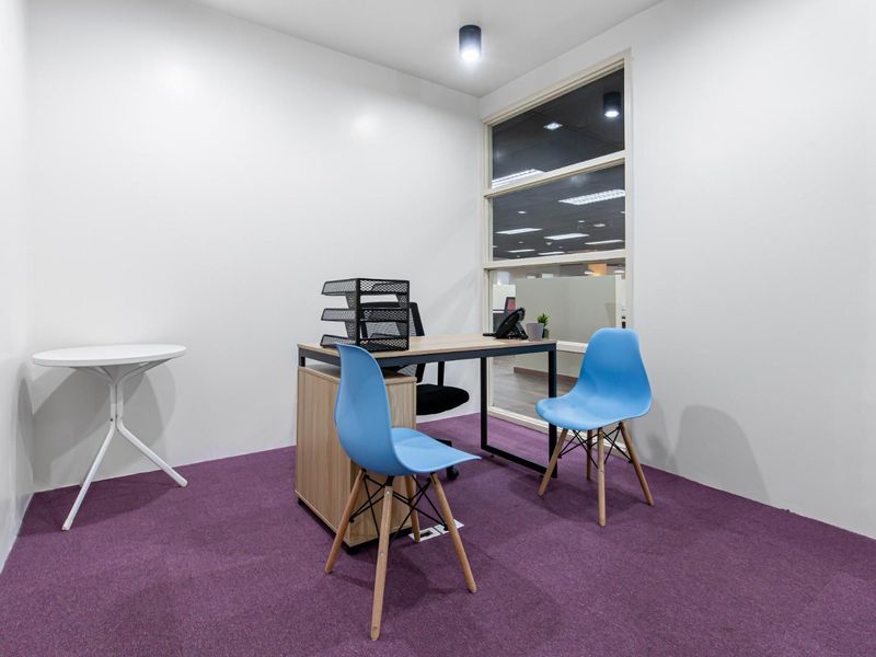 A compact, well-lit office space featuring purple carpeting, white walls, and a large fixed window providing natural light.