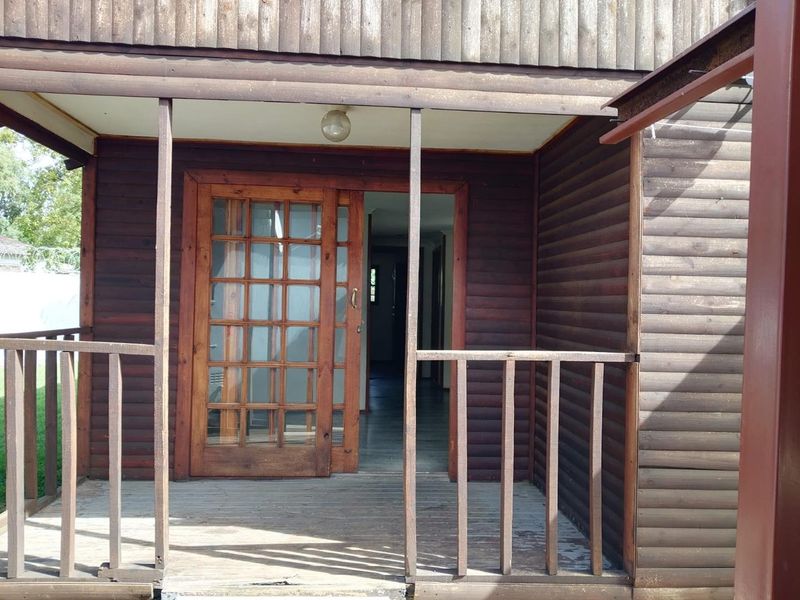 A covered timber patio featuring log-style wall cladding, a multi-pane sliding entrance door, and a wooden balustrade, showing signs of weathering.