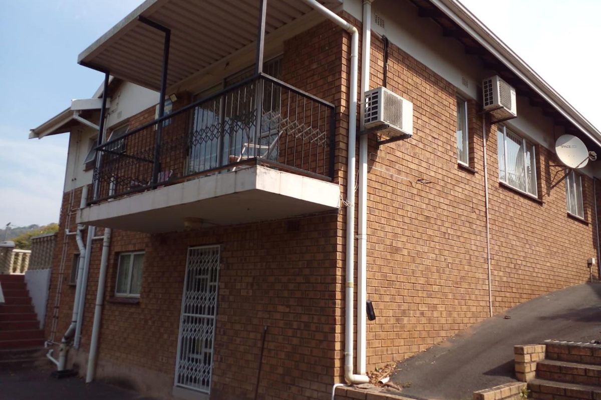Exterior view of a brick house featuring a raised balcony with metal railings, security gates, and external air conditioning units.