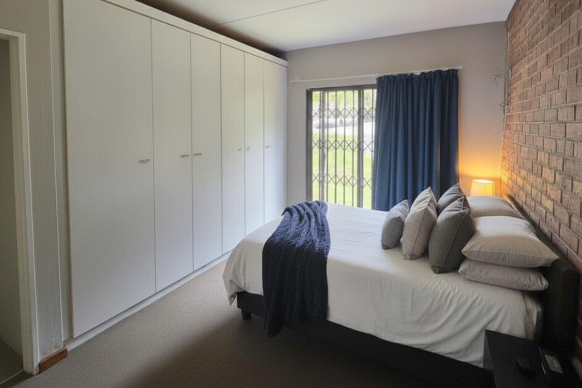 A well-lit bedroom featuring a large wall-to-wall built-in wardrobe, a distinctive exposed brick feature wall, and sliding glass doors with security gate.