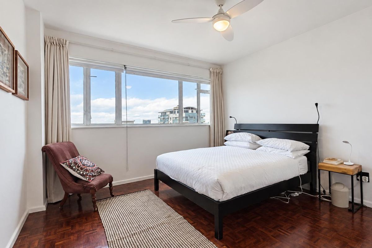 A bright bedroom featuring classic parquet flooring, a ceiling fan, and a large window offering natural light and urban views.