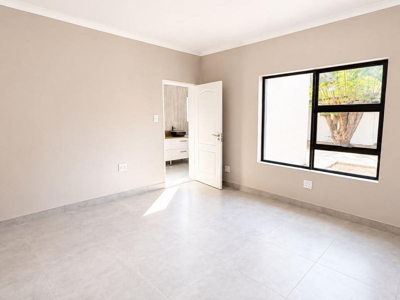 A bright, empty bedroom featuring neutral wall tones, large aluminium-framed windows, and modern tiled flooring, with an adjoining en-suite bathroom visible.