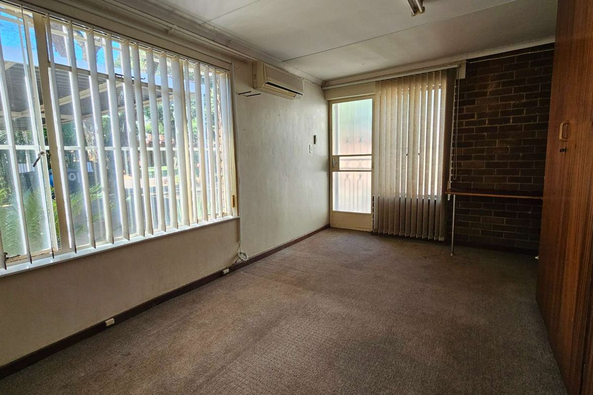 A bedroom featuring built-in wooden cabinetry, a wall-mounted air conditioning unit, and large windows with vertical blinds, offering a blank canvas for renovation.