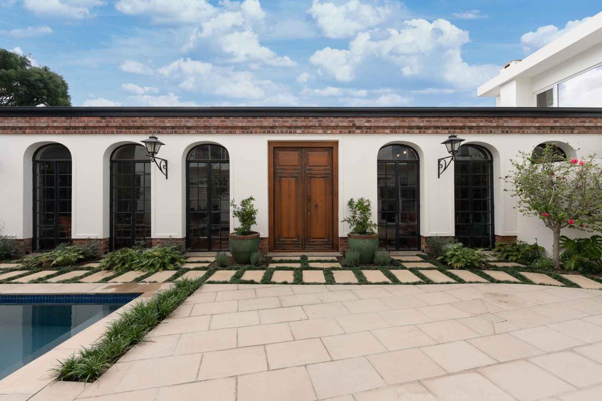 A beautifully maintained rear exterior featuring a swimming pool, stone patio, and elegant arched windows flanking a central timber entrance.