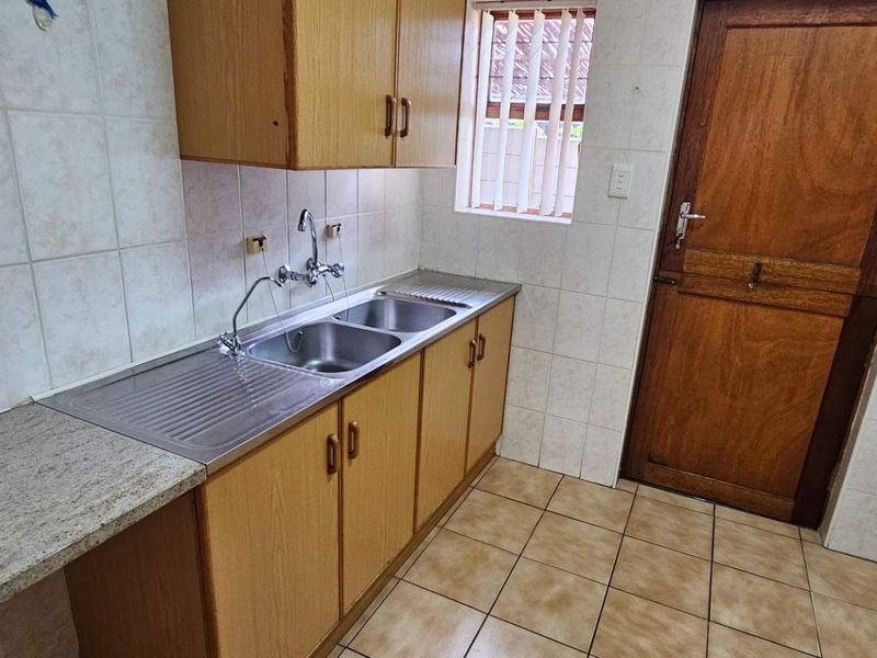 A functional scullery area featuring a double bowl stainless steel sink, laminate cabinetry, tiled splashbacks, and tiled flooring with direct external access.