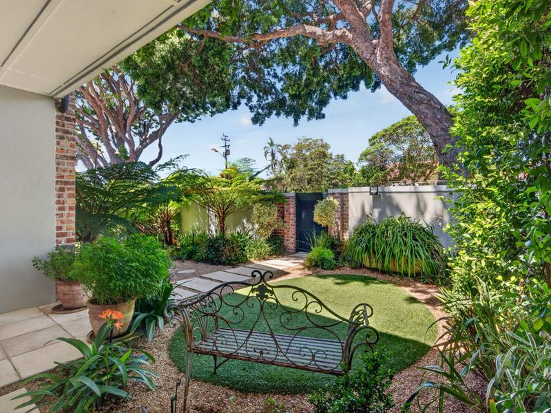 A covered patio area overlooking a landscaped garden with artificial lawn, stone pathways, and a secure boundary wall with a pedestrian gate.