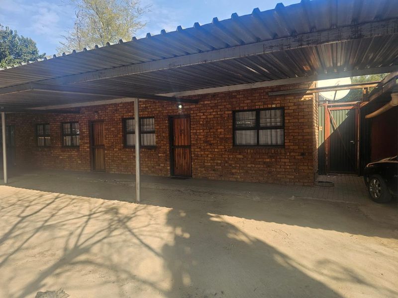 Exterior view of a facebrick building featuring a large corrugated metal carport and secure entrance doors with security gates.