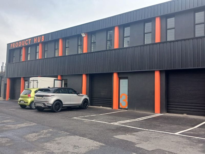 Modern industrial warehouse unit featuring dark metal cladding with orange accents, roller shutter doors, and dedicated asphalt parking bays.