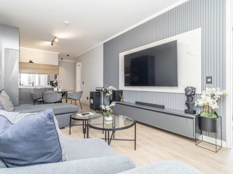 Modern open-plan living area featuring light laminate flooring, a stylish grey feature wall with integrated media unit, and a sleek kitchen in the background.