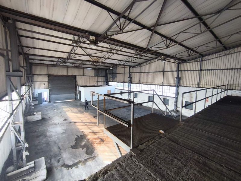 Spacious industrial warehouse featuring a high-clearance steel truss roof, concrete flooring, a roller shutter door, and an elevated mezzanine level with metal access stairs.