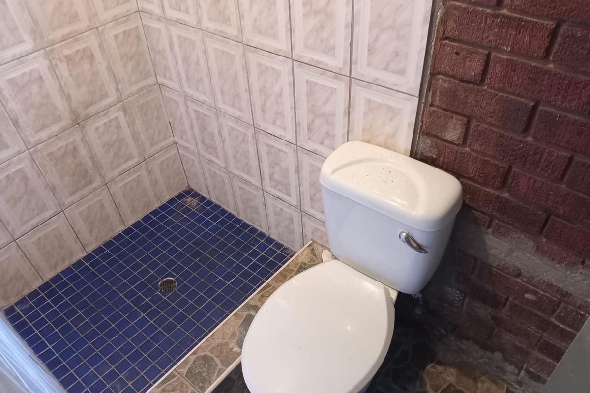 A compact bathroom featuring a toilet and a tiled shower area with exposed brick wall detailing, requiring cosmetic updates to finishes.
