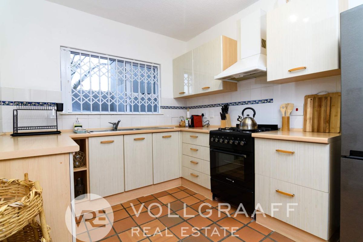 A functional kitchen featuring light-toned cabinetry, a freestanding stove with extractor fan, tiled flooring, and a window with security bars.