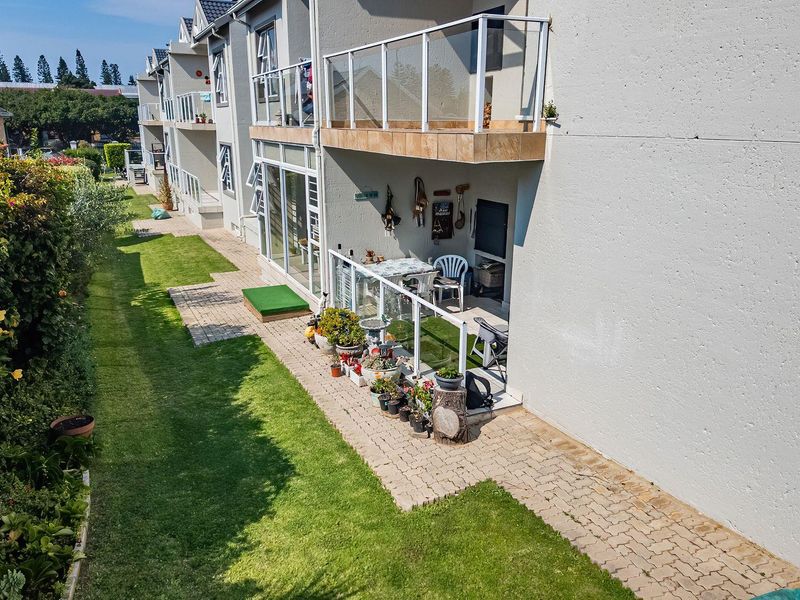 Rear view of the apartment complex featuring ground floor patios with glass balustrades, paved walkways, and well-maintained communal garden areas.
