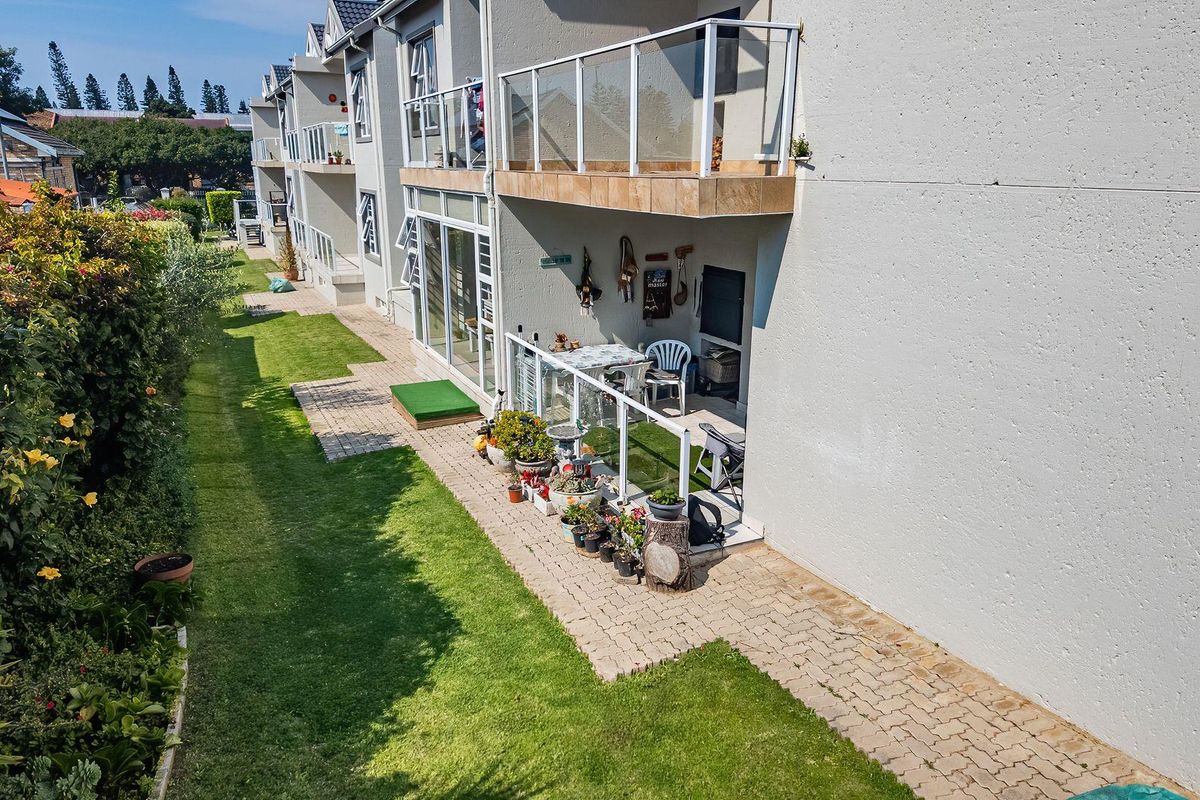 Rear view of the apartment complex featuring ground floor patios with glass balustrades, paved walkways, and well-maintained communal garden areas.