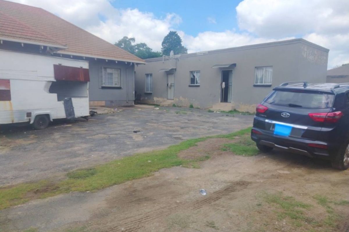 Rear exterior view of the property showing multiple building structures, a paved driveway area, and basic security features on windows and doors.