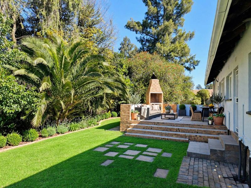A well-maintained rear garden featuring a raised brick patio with a built-in fireplace, lush lawn, and mature landscaping.
