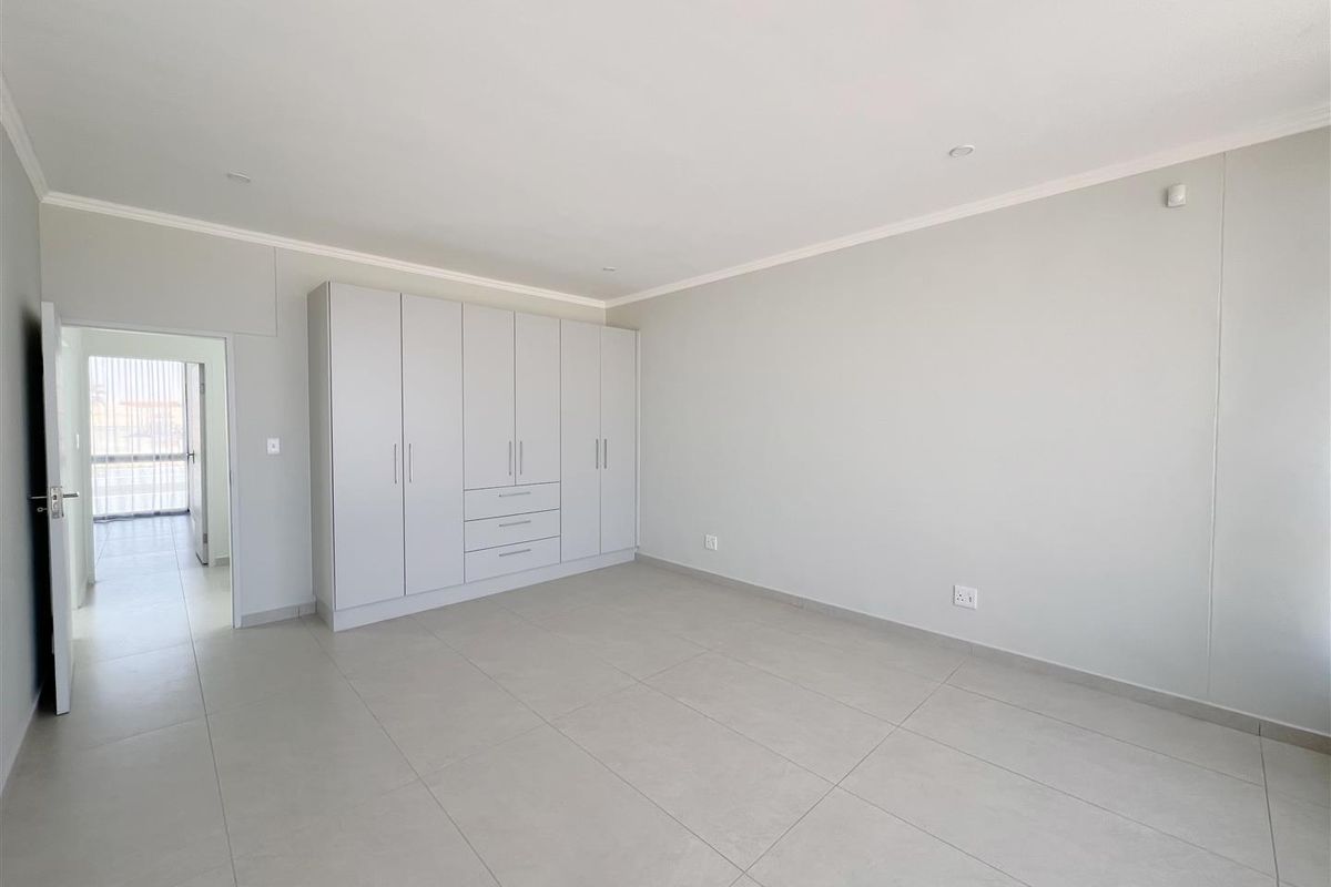 A bright, neutral bedroom featuring large format floor tiles, recessed downlighting, and a substantial built-in wardrobe with ample storage space.
