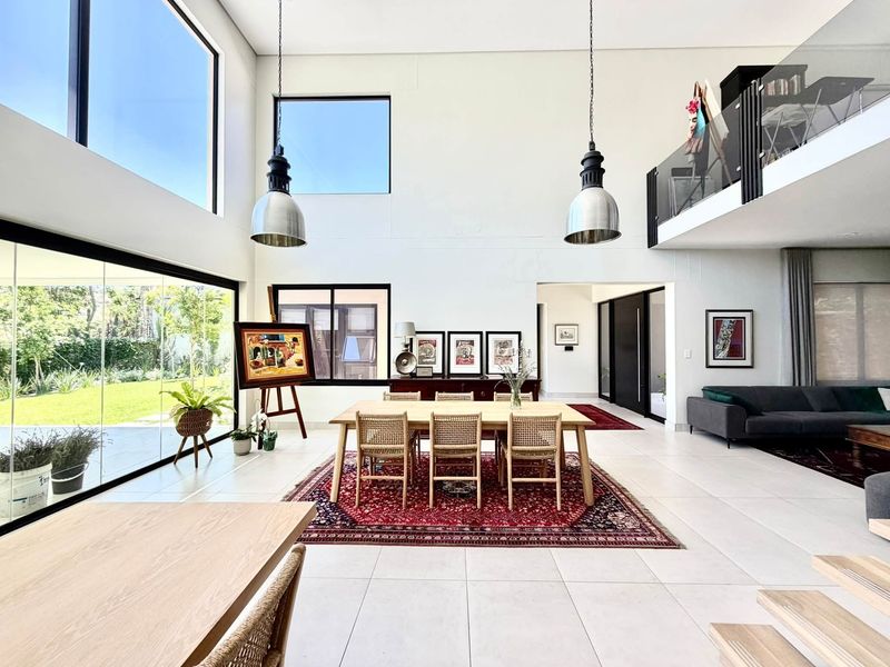Spacious open-plan living and dining area featuring double-volume ceilings, large windows, tiled flooring, and a mezzanine level with a modern glass balustrade.
