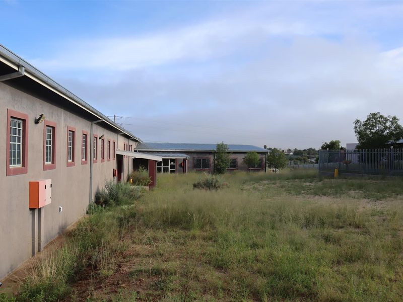 Rear exterior view of the property featuring a long building facade, metal roofing, and an expansive, overgrown yard area with perimeter security fencing.