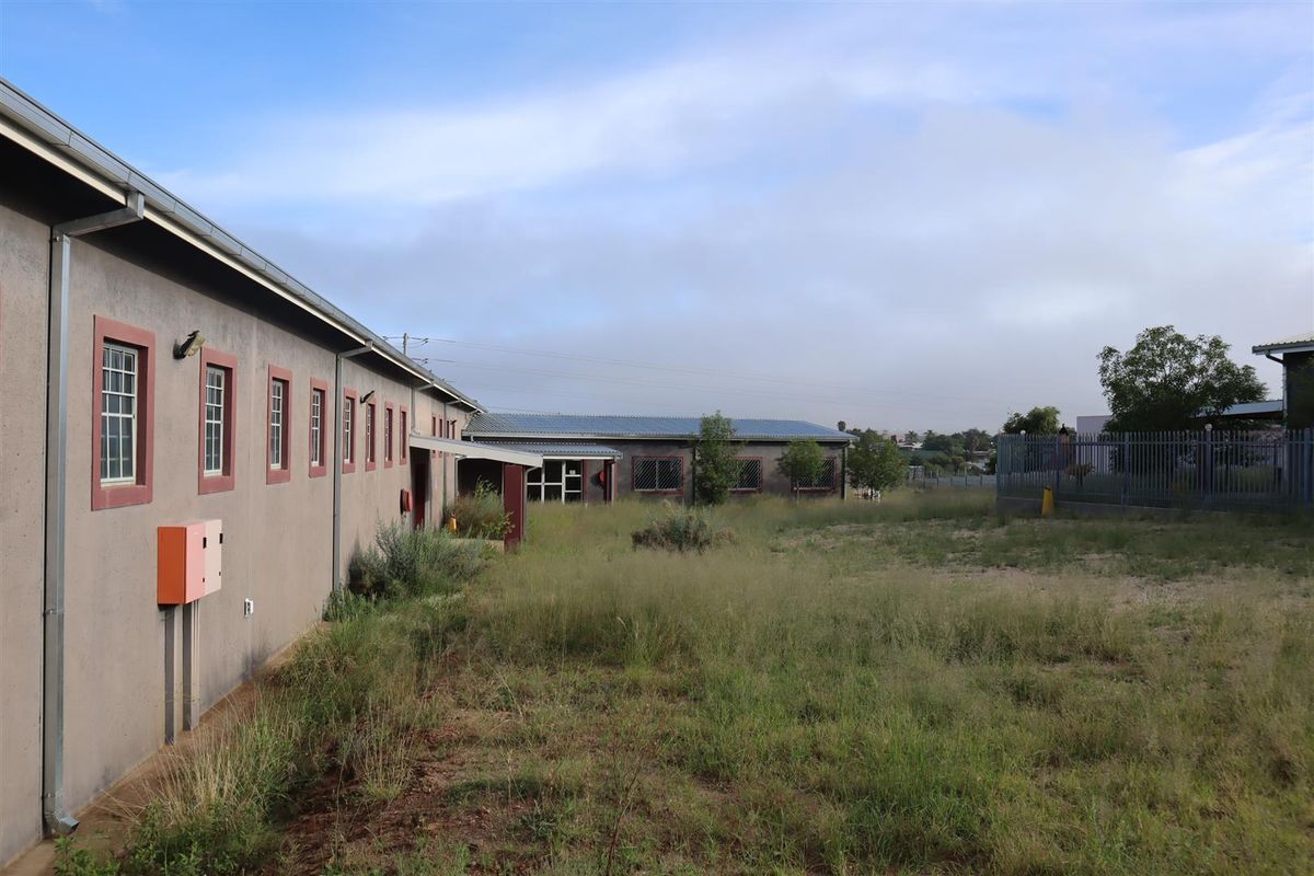 Rear exterior view of the property featuring a long building facade, metal roofing, and an expansive, overgrown yard area with perimeter security fencing.