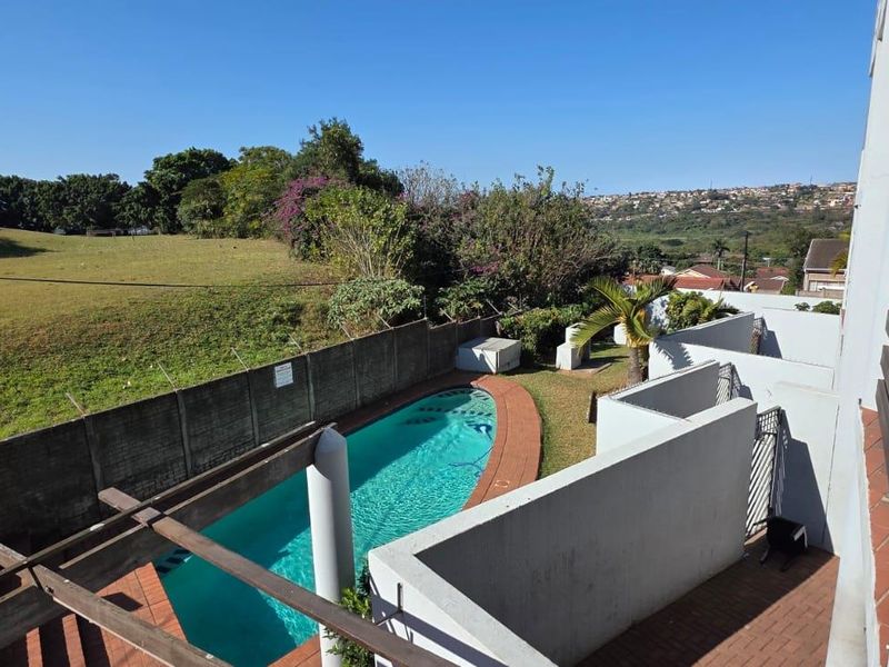 Elevated view of a communal swimming pool area featuring brick paving, a pergola, and a built-in braai, overlooking a grassy landscape.