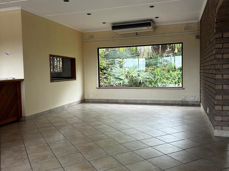 Spacious living area featuring tiled flooring, an exposed brick feature wall, recessed lighting, and a large window overlooking the garden.