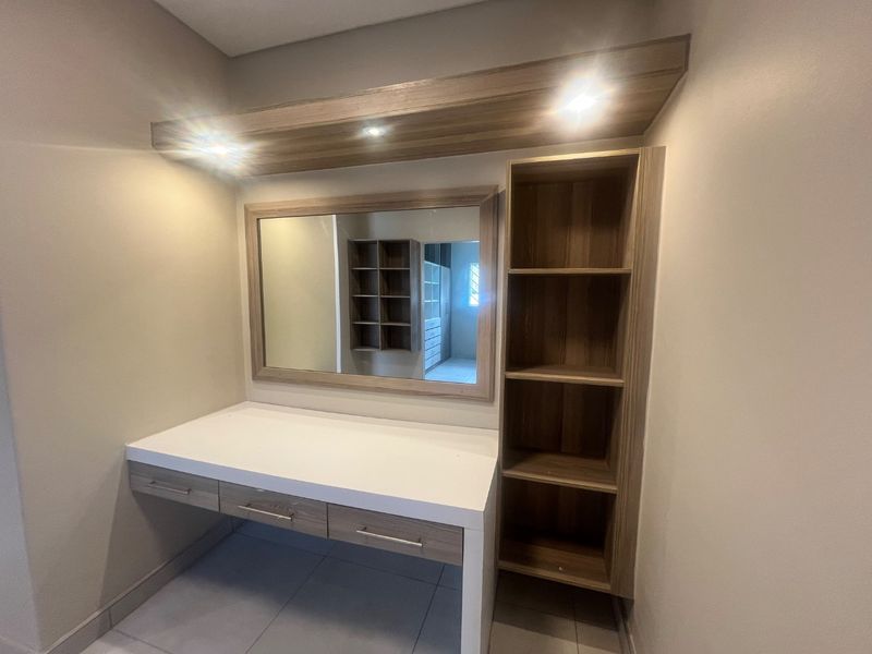 A modern built-in vanity area featuring integrated shelving, a large mirror, and recessed lighting, providing a functional dressing space within the bedroom.