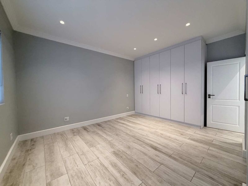 A bright, modern bedroom featuring neutral grey walls, light wood-look laminate flooring, and extensive built-in floor-to-ceiling cupboards.