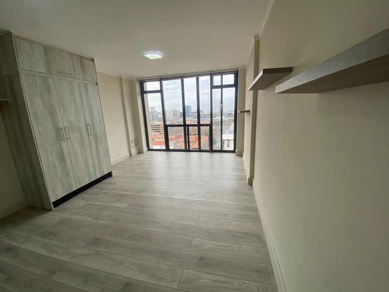 A bright, modern bedroom featuring light wood-look laminate flooring, a built-in wardrobe, floating shelves, and expansive windows offering an urban city view.