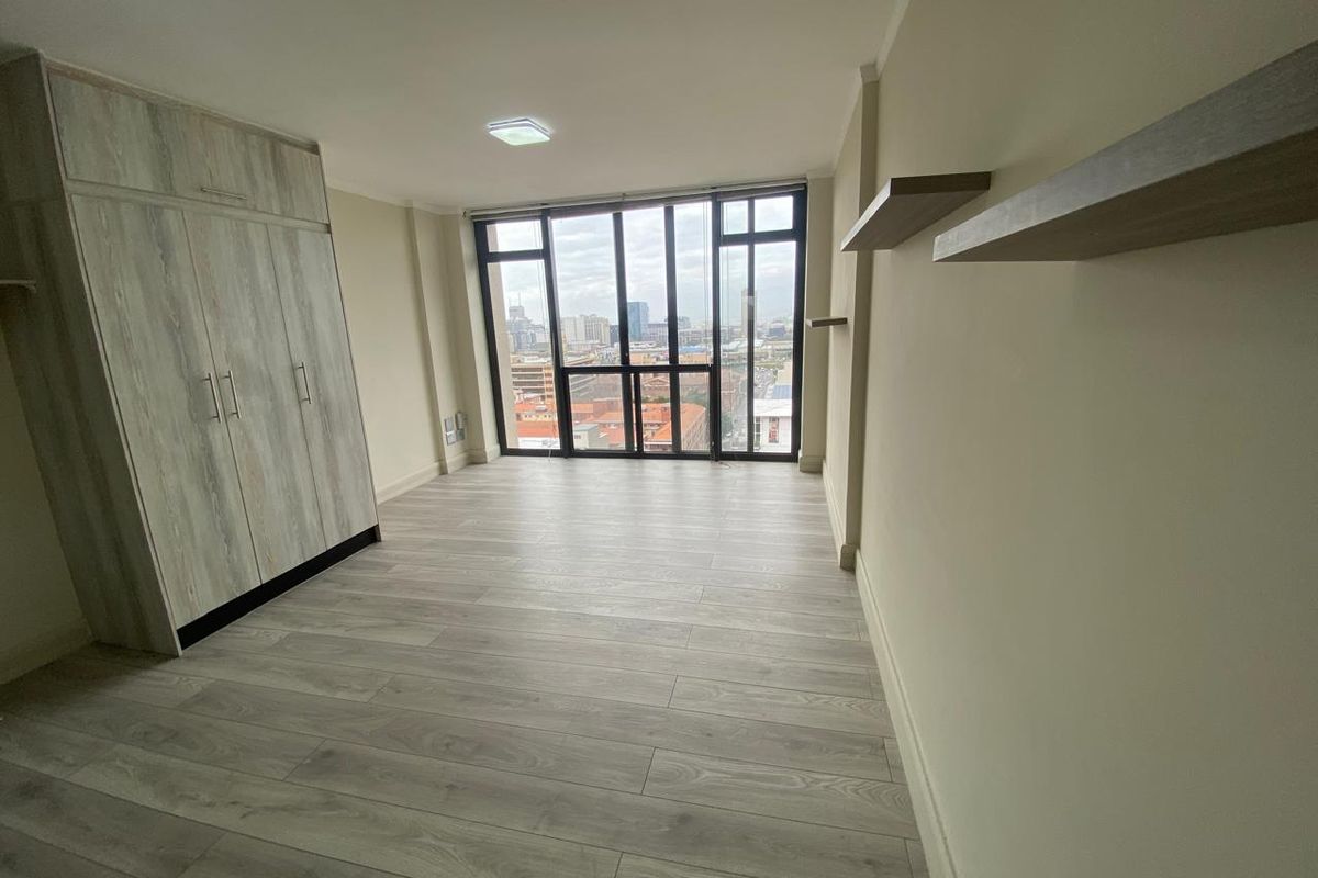A bright, modern bedroom featuring light wood-look laminate flooring, a built-in wardrobe, floating shelves, and expansive windows offering an urban city view.