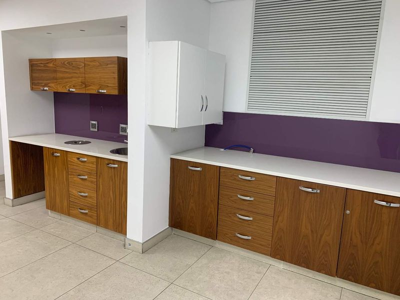 Modern office kitchenette featuring wood-grain cabinetry, white countertops, purple glass splashbacks, and integrated sinks in a clean, well-maintained workspace.