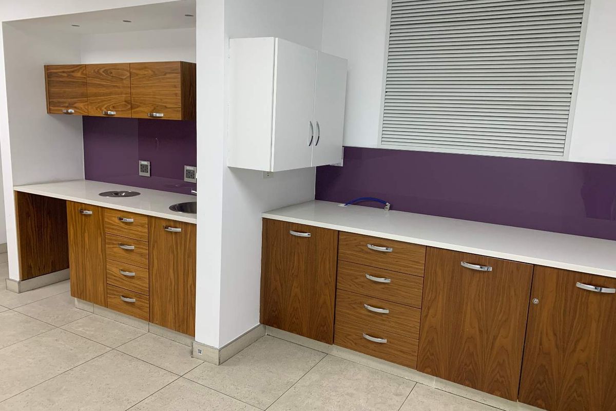Modern office kitchenette featuring wood-grain cabinetry, white countertops, purple glass splashbacks, and integrated sinks in a clean, well-maintained workspace.