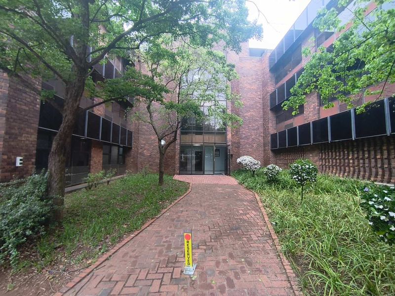 Front entrance of a brick office building featuring a paved walkway, landscaped garden beds, and mature trees leading to a glass-fronted lobby.