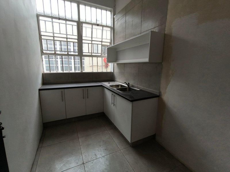 A compact kitchen area featuring l-shaped cabinetry, a stainless steel sink, and industrial-style windows, currently requiring cosmetic wall repairs and updates.