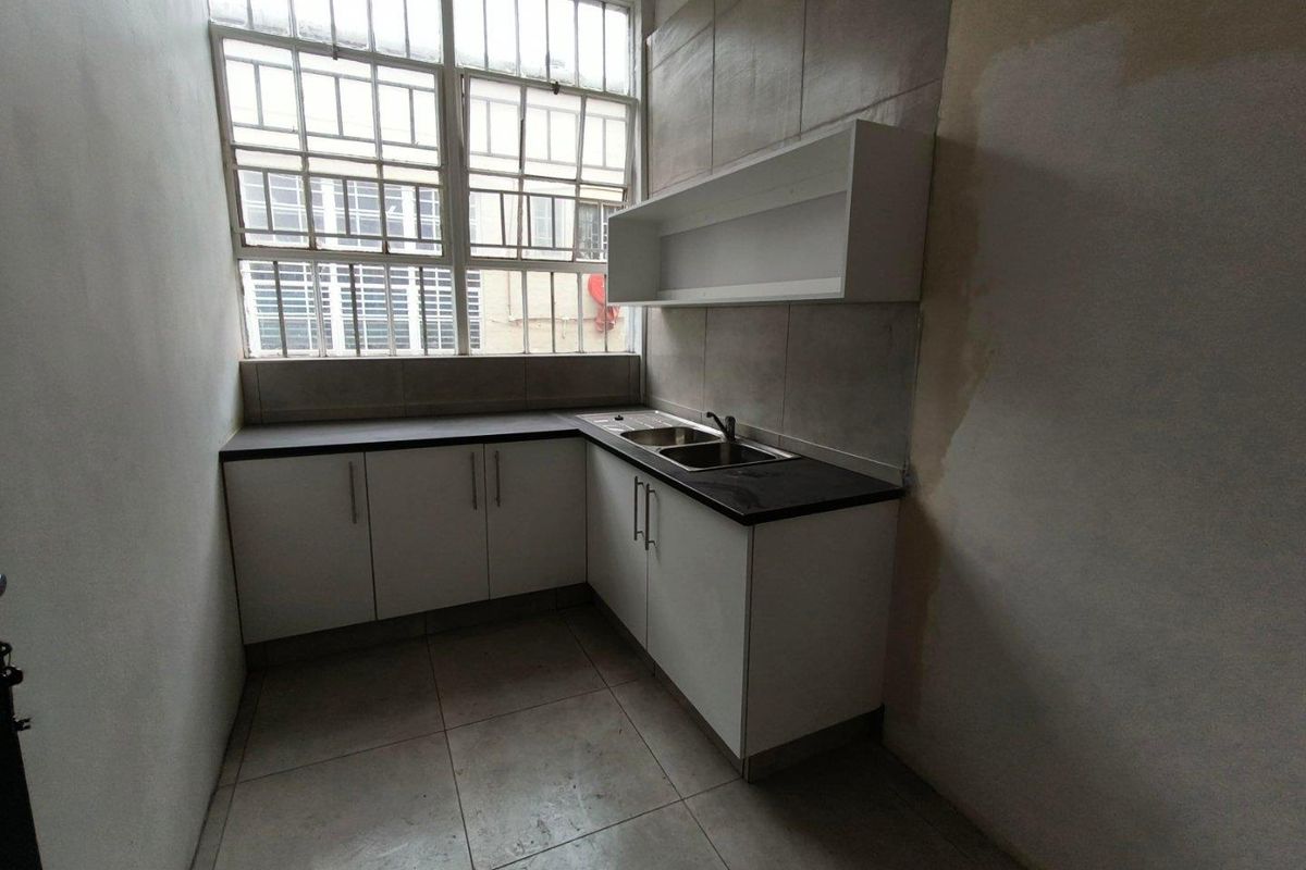 A compact kitchen area featuring l-shaped cabinetry, a stainless steel sink, and industrial-style windows, currently requiring cosmetic wall repairs and updates.