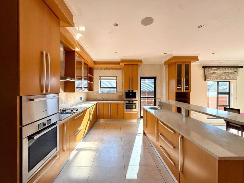 Spacious kitchen featuring warm wood cabinetry, stone countertops, integrated appliances, and a large breakfast bar island with tiled flooring.