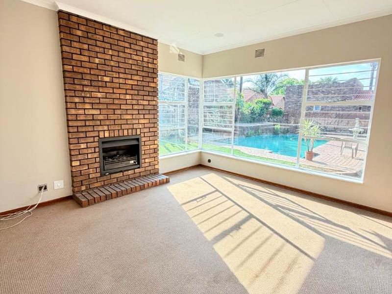 Spacious living room featuring a classic brick fireplace and large corner windows offering views of the swimming pool area.