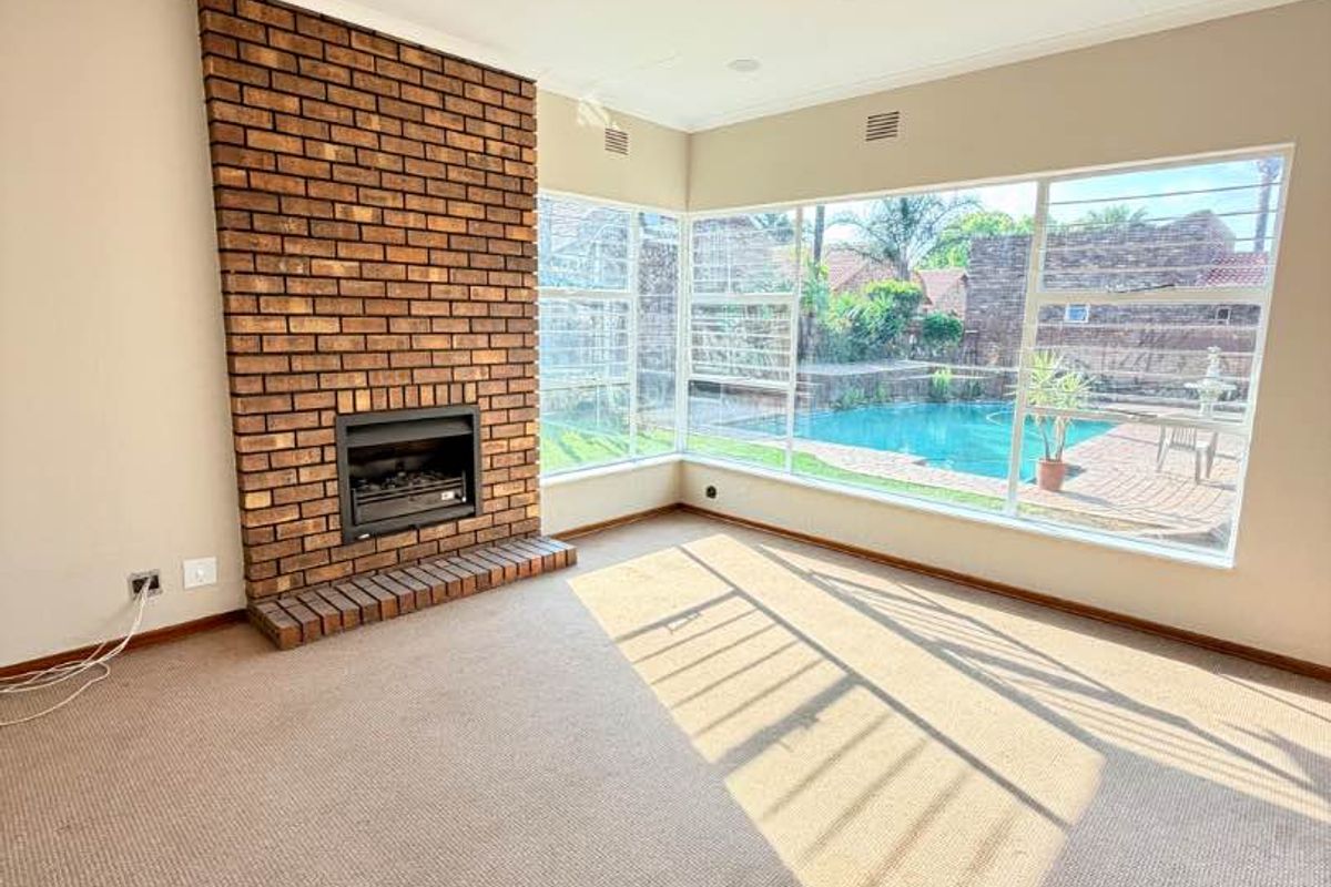 Spacious living room featuring a classic brick fireplace and large corner windows offering views of the swimming pool area.