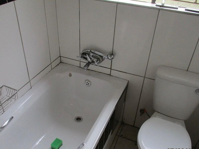 A functional bathroom featuring a standard bathtub with a mixer tap and a toilet, finished with white wall tiles and tiled flooring.