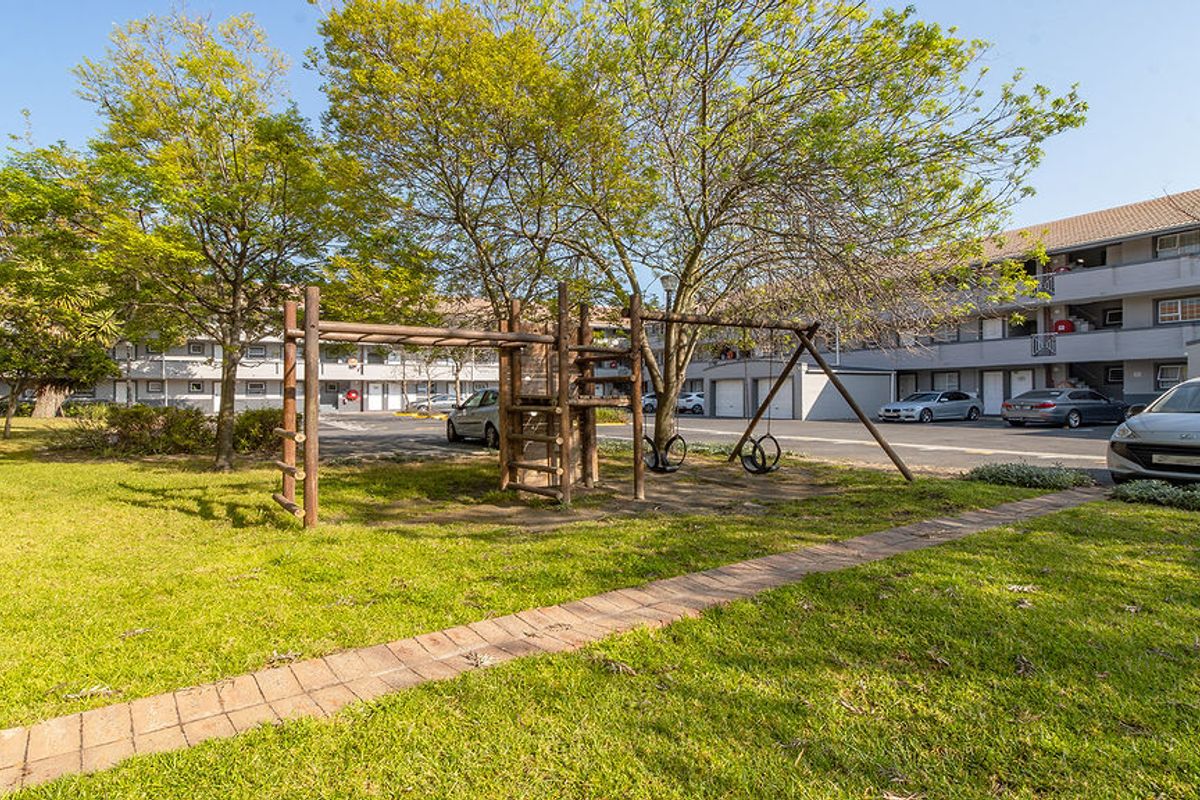 A view of the apartment complex exterior featuring a communal garden, paved walkway, and playground area with adjacent parking facilities.