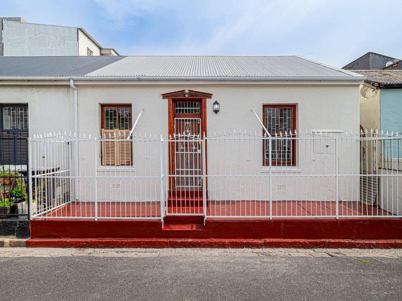 Front facade of a charming cottage featuring a corrugated metal roof, secure wrought iron fencing, and traditional timber window and door frames.