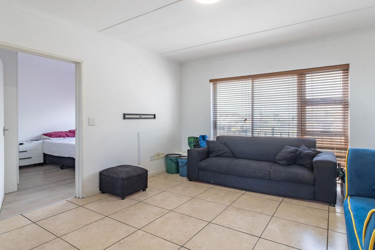 A living room featuring neutral tiled flooring, white walls, and venetian blinds, with an open doorway leading to an adjacent bedroom.