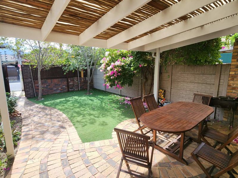 A covered outdoor patio area featuring a timber pergola, brick paving, and a well-maintained artificial lawn with established garden borders.