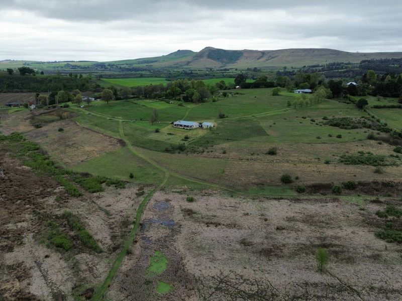 An aerial view of a large, open residential vacant land plot featuring rolling green pastures and a small existing dwelling in the distance.