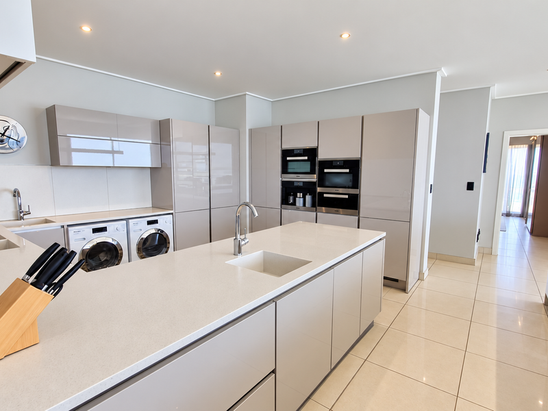 Modern kitchen featuring high-gloss cabinetry, stone countertops, a central island with sink, and integrated appliances including oven, microwave, and laundry units.