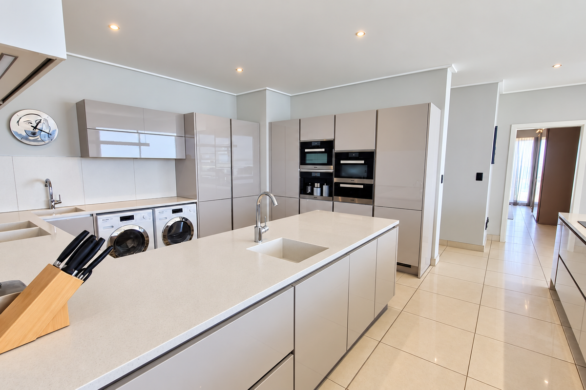 Modern kitchen featuring high-gloss cabinetry, stone countertops, a central island with sink, and integrated appliances including oven, microwave, and laundry units.