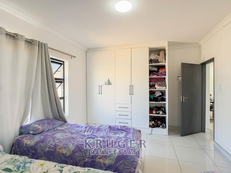 A well-lit bedroom featuring neutral tiled flooring, white built-in cupboards with integrated shelving, and a large window with curtain fixtures.